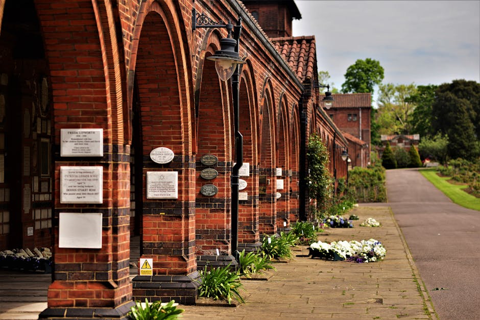 The image shows a row of red-brick buildings with arched windows and multiple black lantern-style lamps mounted along the wall. Along the paved walkway outside the buildings, there are fresh floral bo