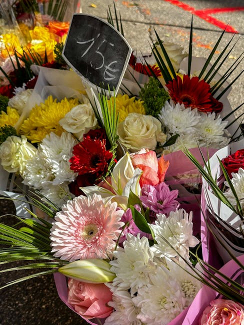 A colorful floral bouquet featuring a mix of white roses, pink gerbera daisies, white lisianthus, red gerbera daisies, and paler pink roses, arranged with long green palm leaves and other foliage. The