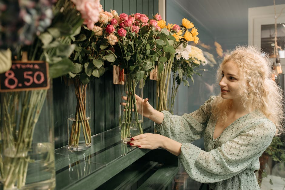 A woman with curly blonde hair and a light floral blouse arranging a bouquet of fresh flowers in a glass vase at a flower shop. The bouquet includes pink roses, yellow chrysanthemums, and white flower