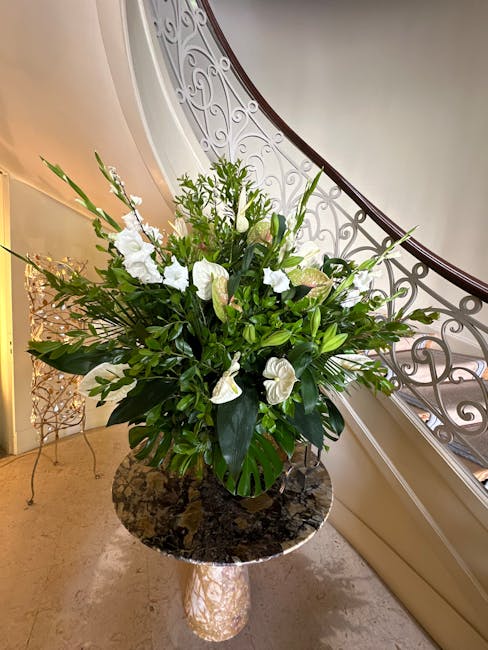 A tall, lush floral arrangement featuring predominantly white flowers, including large calla lilies and roses, complemented by assorted green foliage such as glossy leaves and ferns. The bouquet is pl