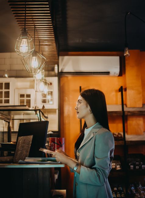The image shows a woman with long dark hair and glasses, dressed in a light grey blazer, standing at a counter inside a modern, warmly lit space. She is holding a tablet in her right hand and appears 