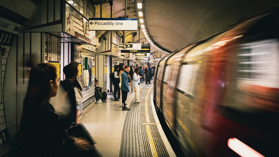 A lively London Underground platform at Pinner station, with passengers waiting on the tiled floor and a blurred train arriving or departing on the right. The station has striped black and white wall 