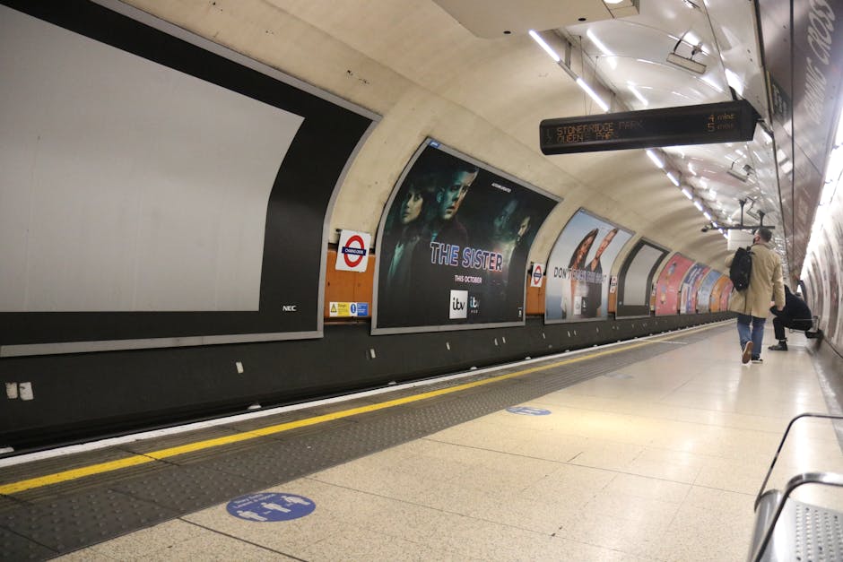 A photograph of an underground station platform showing a curved, beige ceiling with modern lighting fixtures. The platform features advertising posters, including one for the film 'The Sister,' displ