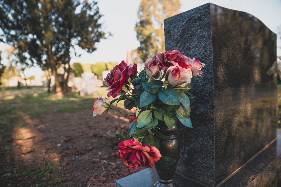 A floral arrangement featuring pink and red roses with hints of white, placed in a small black vase on a polished surface next to a dark granite gravestone. The roses are fresh with slightly ruffled p