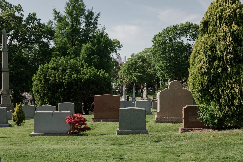 A peaceful cemetery scene showing several upright headstones made of granite and marble in varying shades of grey, brown, and black, arranged across a well-maintained green lawn. In the foreground, a 