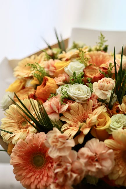 A vibrant floral arrangement featuring a mix of blush pink gerbera daisies, soft peach roses, white lisianthus, and pale yellow ranunculus, all accented with sprigs of green foliage and tall, slender 