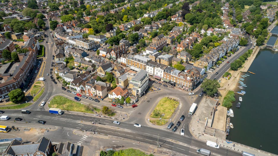 Aerial view of the Pinner town centre with a cluster of Victorian and modern buildings, streets with cars and pedestrians, and a river with moored boats on the right side. The scene shows lush green t