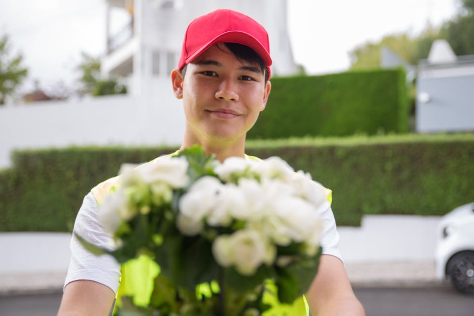 A smiling delivery person wearing a red baseball cap and a neon yellow vest is holding a bouquet of fresh white flowers, including roses and other blossoms, wrapped in clear cellophane and tied with a