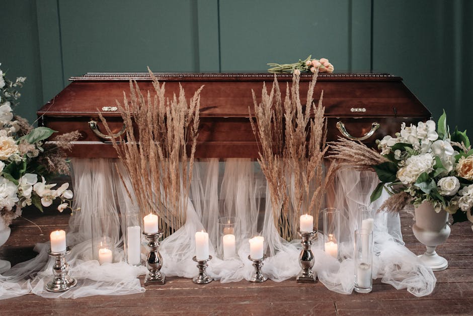 A floral arrangement featuring white roses, orchids, and pale pink blooms in white ceramic vases on either side of a wooden casket, decorated with dried pampas grass and soft white fabric draping down