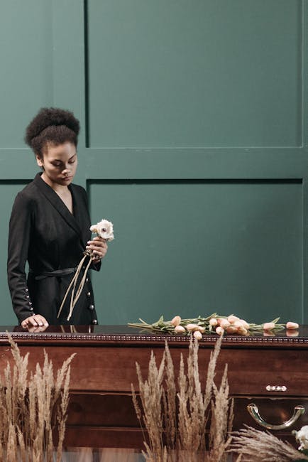 A young woman with dark skin and natural curly hair, dressed in a black outfit, is holding a small white and pink floral arrangement in her right hand. She stands beside a wooden casket draped with a 