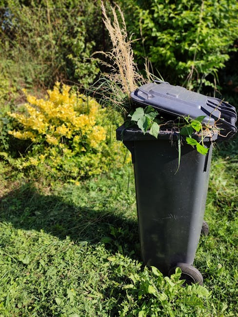 A black wheelie bin placed outdoors on a grassy area, filled with discarded floral arrangements consisting of various fresh flowers such as yellow, pink, and white blooms, as well as greenery and pamp