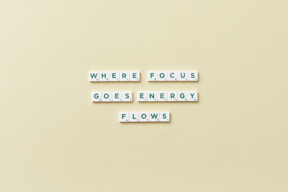 A flat lay of white scrabble tiles arranged on a pale yellow background spell out the phrase 'WHERE FOCUS GOES ENERGY FLOWS', with each word on a separate line. The tiles are neatly aligned, featuring